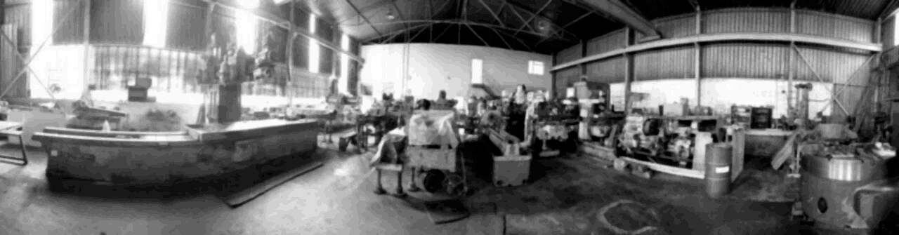 A long machine bed in the foreground of a workshop floor, with a row of machines under a corrugated roof behind it.